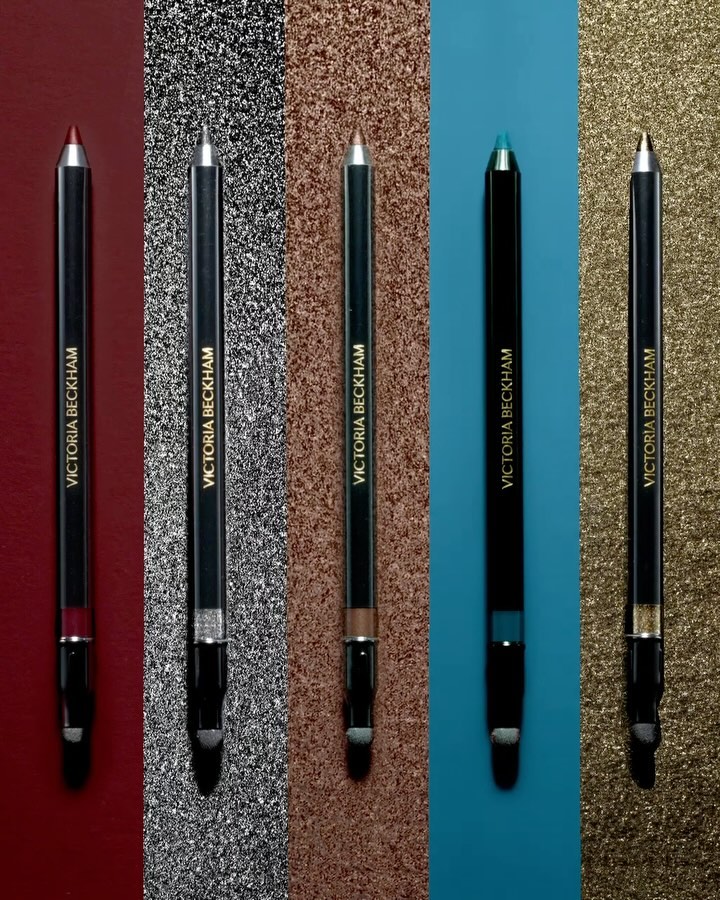 Five Victoria Beckham eyeliner pencils in various colors arranged against shimmering backgrounds for a beauty product shot.