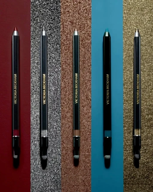 Five Victoria Beckham eyeliner pencils in various colors arranged against shimmering backgrounds for a beauty product shot.