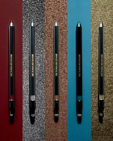 Five Victoria Beckham eyeliner pencils in various colors arranged against shimmering backgrounds for a beauty product shot.