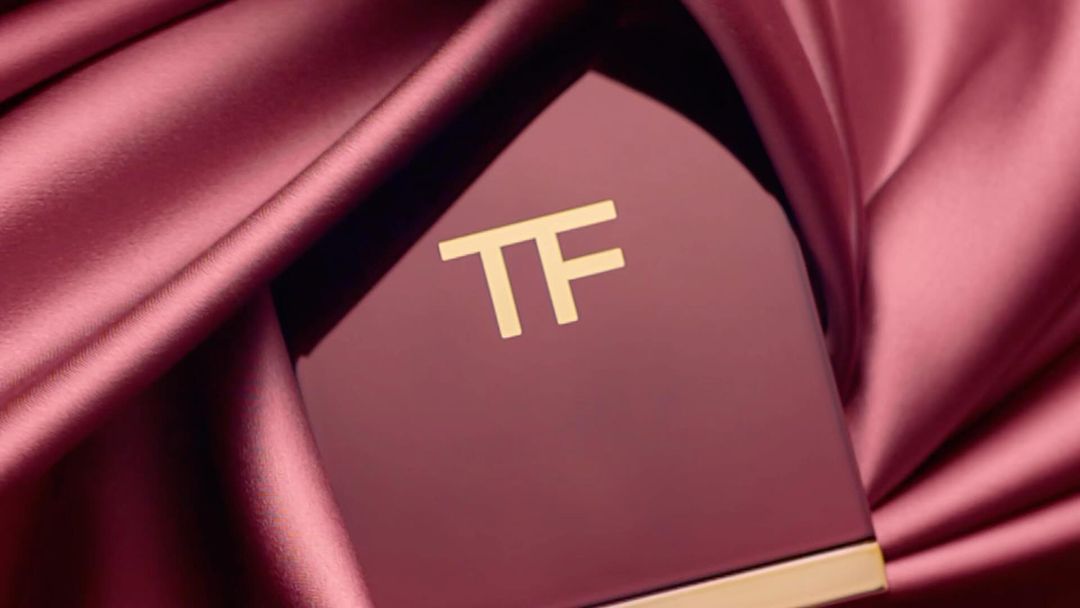 Close-up of a burgundy Tom Ford beauty compact with gold logo on a burgundy satin background. Beauty product shot.