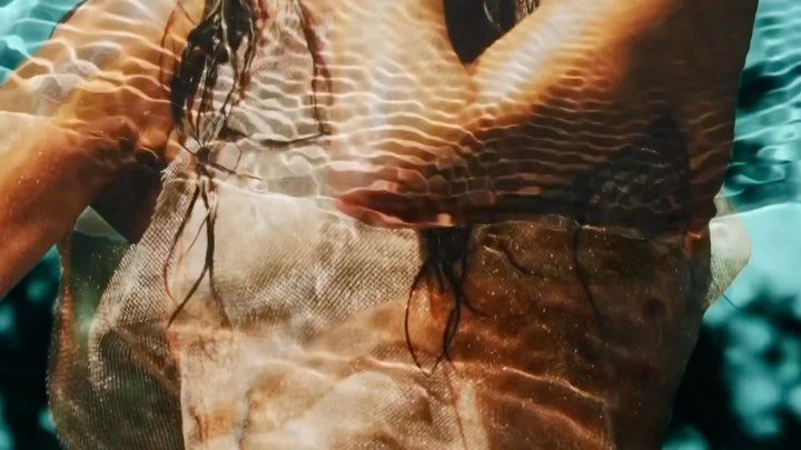 Underwater view of a person with wet hair and a light colored top with water patterns and teal water background.