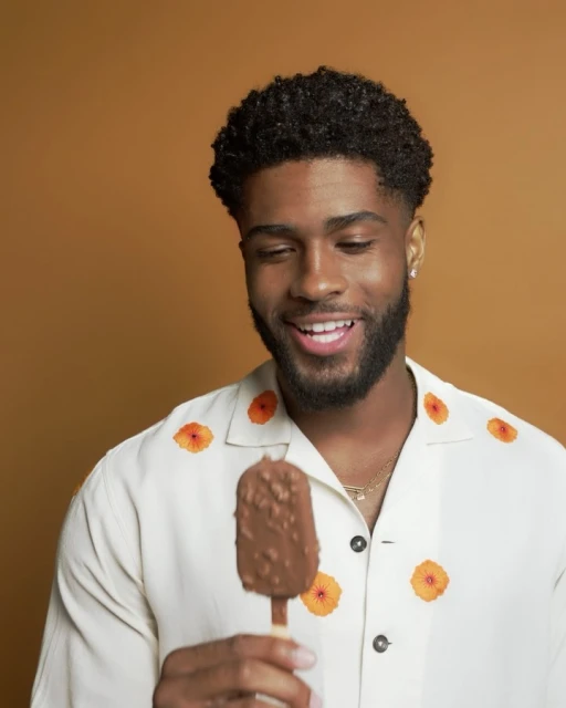 Cheerful Black man smiles while holding a chocolate ice cream bar. He wears a white shirt with orange floral pattern against a tan backdrop.