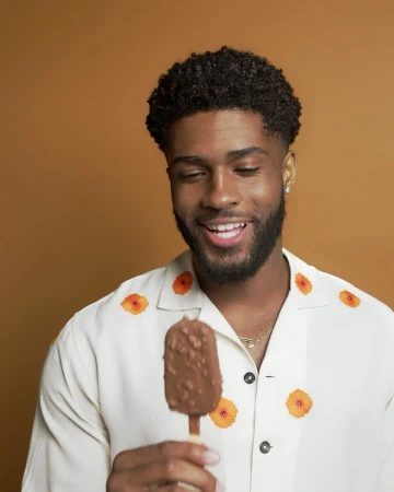 Cheerful Black man smiles while holding a chocolate ice cream bar. He wears a white shirt with orange floral pattern against a tan backdrop.