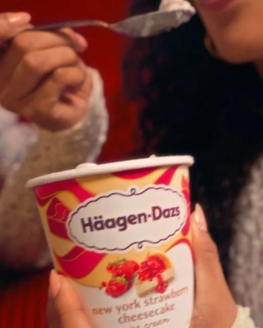 A person is holding a container of Häagen-Dazs New York Strawberry Cheesecake ice cream and eating some with a spoon.