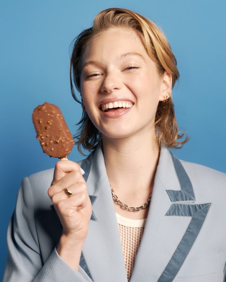 Smiling woman in blue blazer holds a chocolate-covered ice cream bar against a blue background. Portrait shot, bright studio lighting.