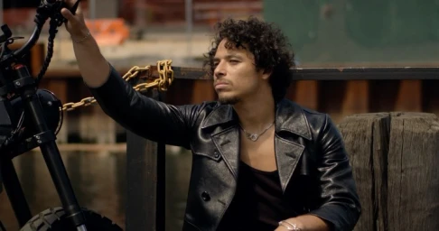 Man with curly hair wearing a black leather jacket seated next to a motorcycle near a waterfront.