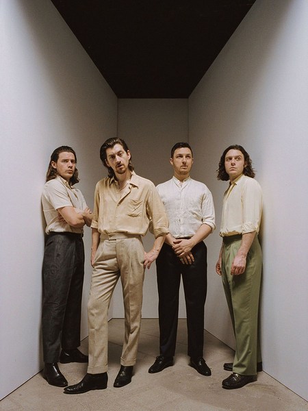 Indoor portrait of four adult men in a minimalist setting, wearing shirts and pants in shades of beige, white, green, and black.