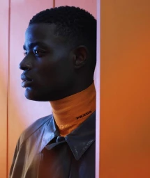 Close-up profile of a black man wearing an orange turtleneck and dark leather jacket against an orange wall.