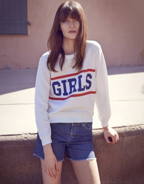 An adult female model with long brown hair wears a white 'GIRLS' sweatshirt and denim shorts, posing against a pink wall outdoors.
