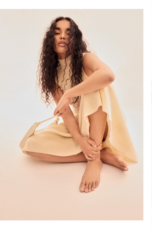 Woman with long curly hair sits in a knitted dress and holds a bag in a light studio setting. Fashion, style, and beauty.
