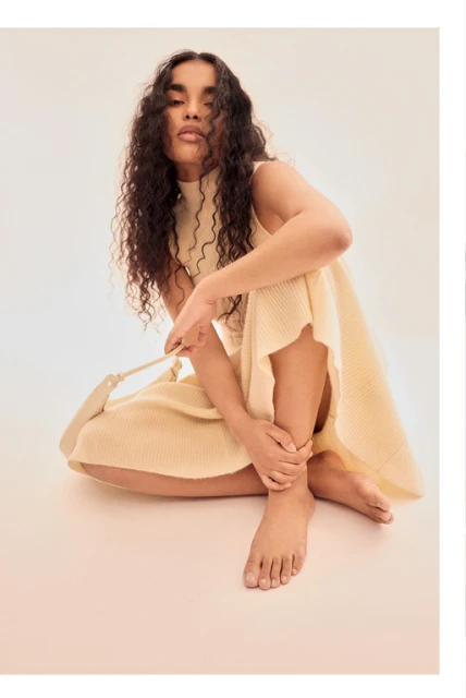 Woman with long curly hair sits in a knitted dress and holds a bag in a light studio setting. Fashion, style, and beauty.