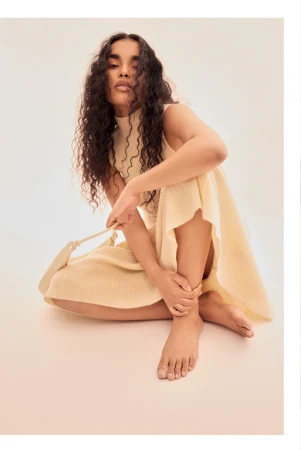 Woman with long curly hair sits in a knitted dress and holds a bag in a light studio setting. Fashion, style, and beauty.