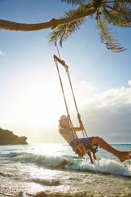 Adult female enjoys a swing hanging from a palm tree on a beach at the ocean's edge with bright sunlight and waves, a tropical getaway.