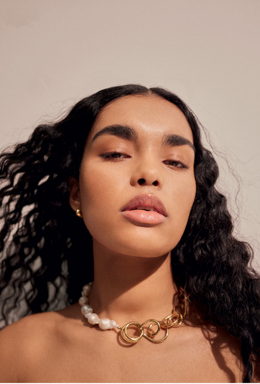 Close-up portrait of a woman with long dark wavy hair wearing a statement pearl and gold chain necklace against a light background.
