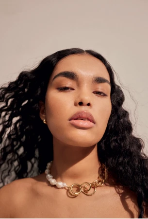 Close-up portrait of a woman with long dark wavy hair wearing a statement pearl and gold chain necklace against a light background.