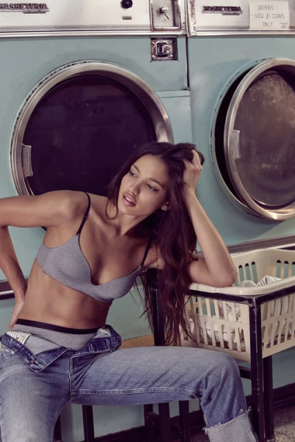 A brunette woman in a grey bra and blue jeans poses sitting near washing machines in a laundromat, looking off to the side.