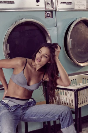 A brunette woman in a grey bra and blue jeans poses sitting near washing machines in a laundromat, looking off to the side.