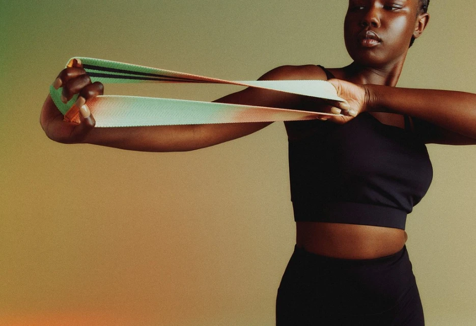 Athletic woman with dark skin in black activewear pulls green resistance band straight, against warm gradient background in medium shot.