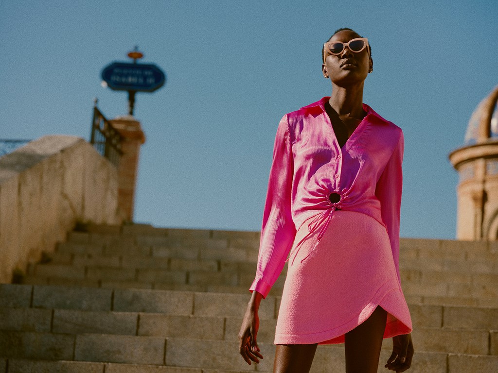 Fashionable woman in pink outfit poses on stone steps against clear sky. Stylish long-sleeve top, skirt, sunglasses, and jewelry.