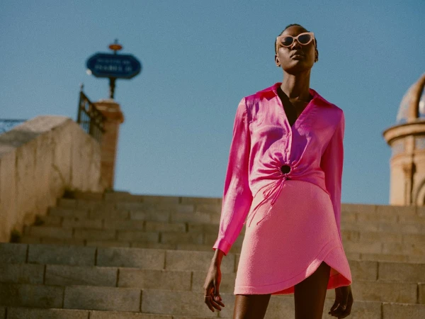 Fashionable woman in pink outfit poses on stone steps against clear sky. Stylish long-sleeve top, skirt, sunglasses, and jewelry.