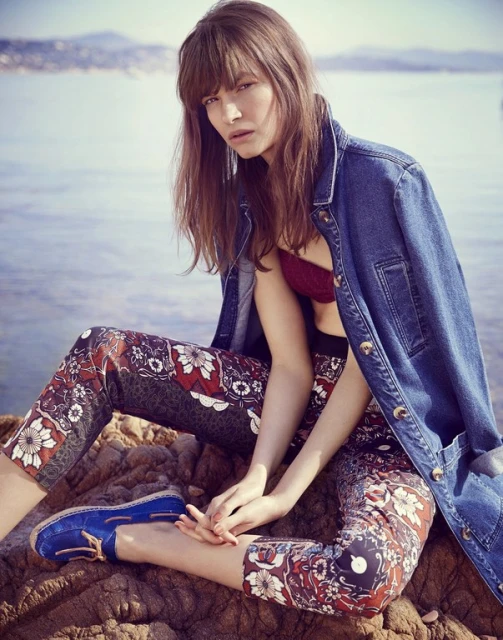 Fashion portrait of a woman with long brown hair wearing a denim jacket and patterned pants, sitting on rocks by the water.