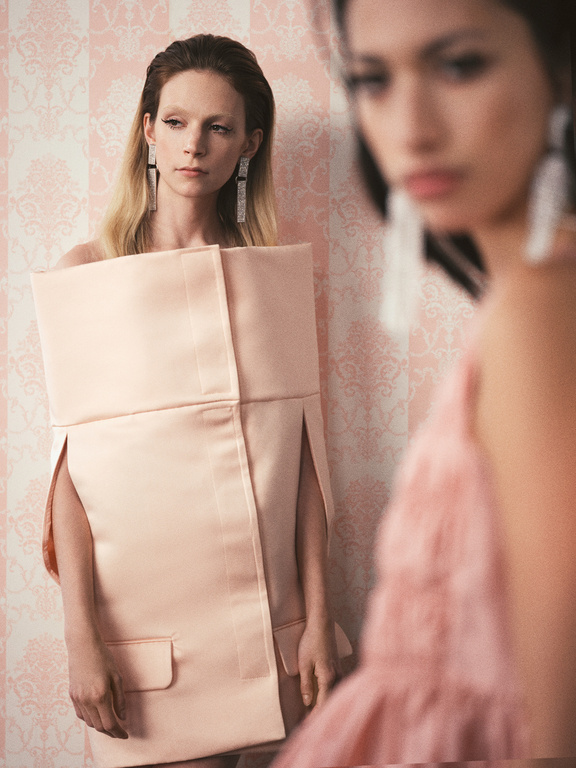 Two women in a photo studio. One stands in the foreground wearing a modern, boxy dress, and the other is blurred in the background.