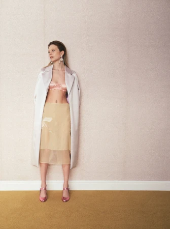 Woman in lingerie and long coat poses against a textured wall with a blonde wig and pink shoes in a studio setting.