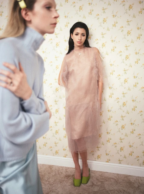 Two women in an indoor fashion shot. One woman wears a blue sweater, and the other wears a light pink dress. Floral wallpaper backdrop.