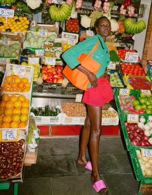 Woman poses in turquoise cardigan and red shorts at a produce stand with colorful fruits and vegetables in the background.