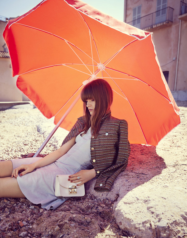 Woman in jacket and dress sits near orange umbrella on gravel with building in the background, holding purse, sunny day.