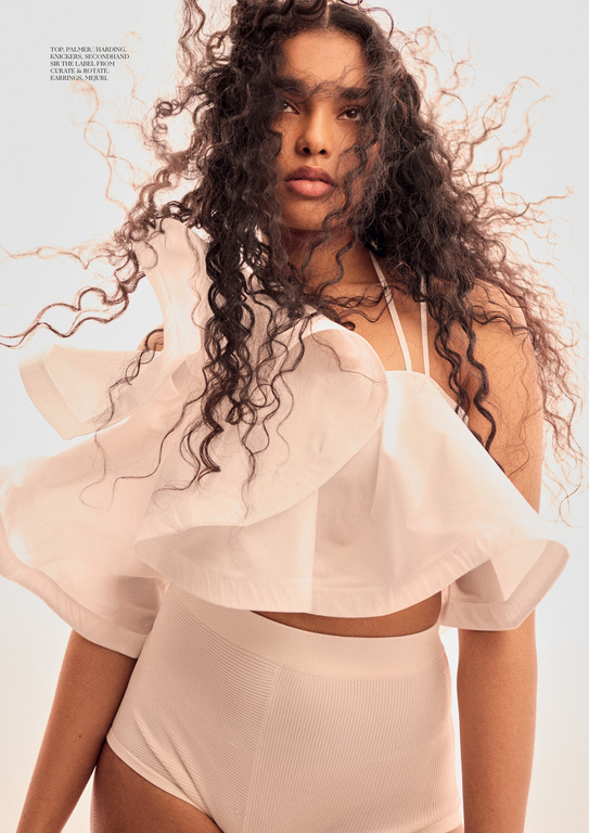 Fashion portrait of an East Indian woman with dark curly hair wearing a light pink ruffled crop top and matching bottoms in a studio.