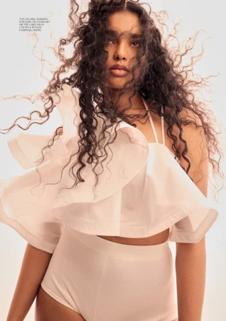 Fashion portrait of an East Indian woman with dark curly hair wearing a light pink ruffled crop top and matching bottoms in a studio.