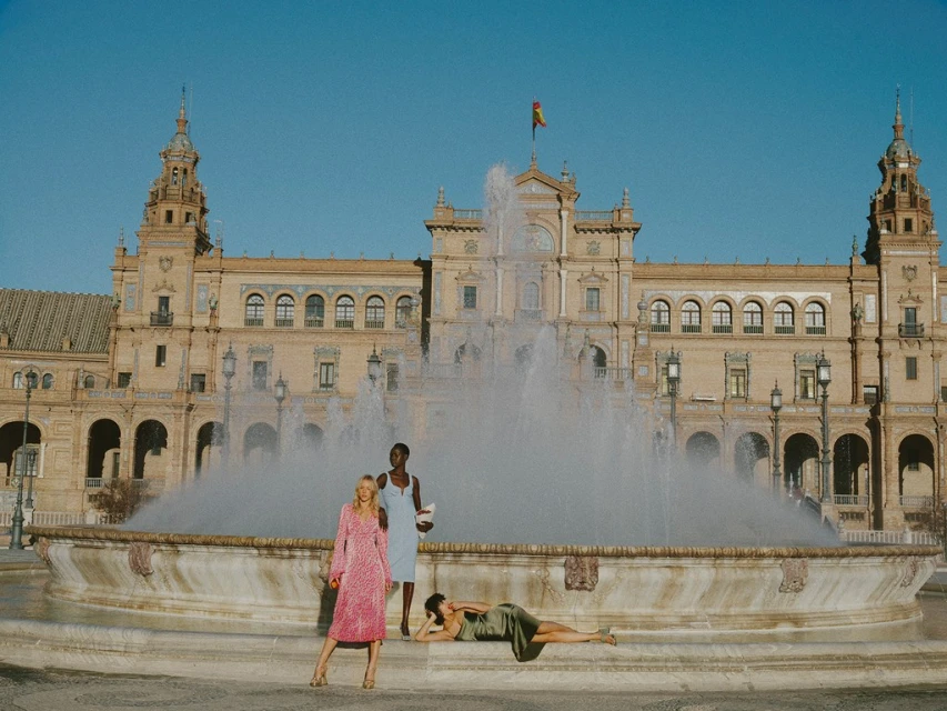 Three women pose near a fountain in Seville, Spain, with a historic building and clear sky in the background, showcasing fashion apparel.