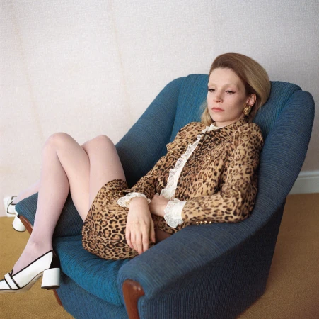 Woman in leopard-print suit lounges in blue armchair, accessorized with white heels and gold earrings in an indoor setting.