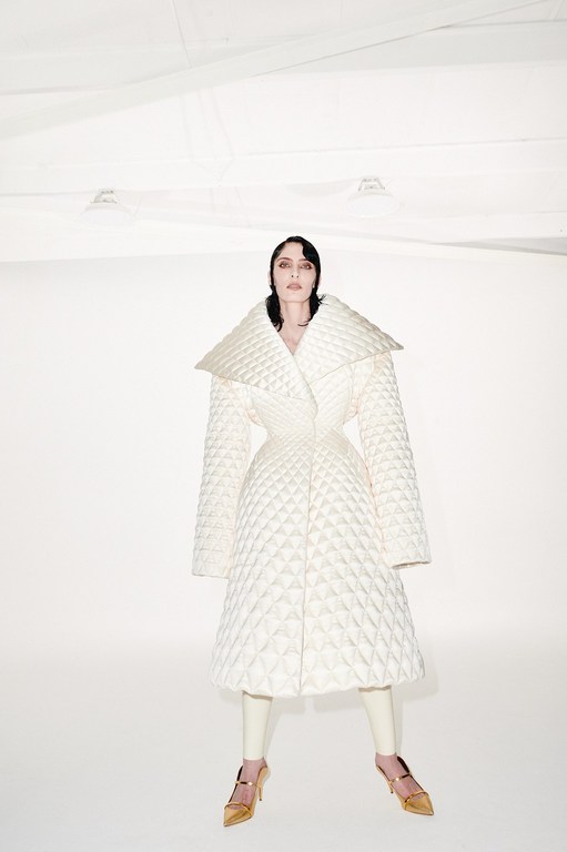 A fashion shot of a woman in a white quilted coat with gold shoes, standing in a bright, stark white studio environment.
