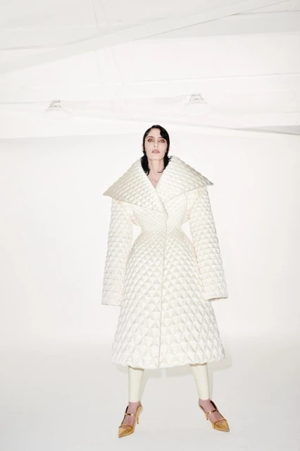 A fashion shot of a woman in a white quilted coat with gold shoes, standing in a bright, stark white studio environment.