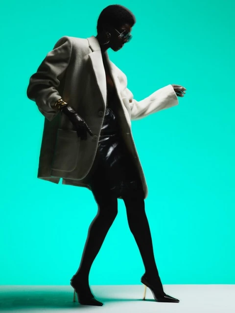 Stylish African-American woman in black skirt, jacket, heels and sunglasses posing against a vibrant teal background.