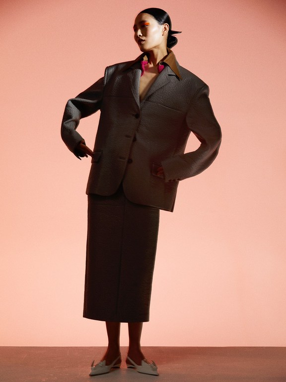 A woman in a gray suit stands in front of a peach-colored backdrop, looking off to the side with a neutral expression in a studio setting.