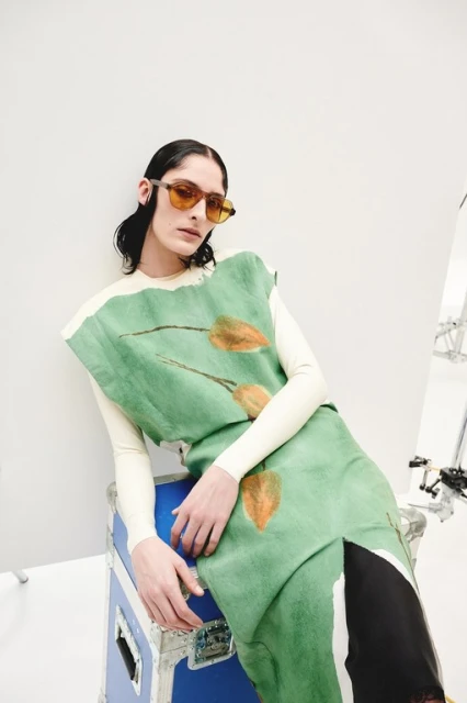 A person with dark hair poses indoors wearing sunglasses, a sleeveless green patterned top and a long-sleeved cream top leaning on a blue road case.