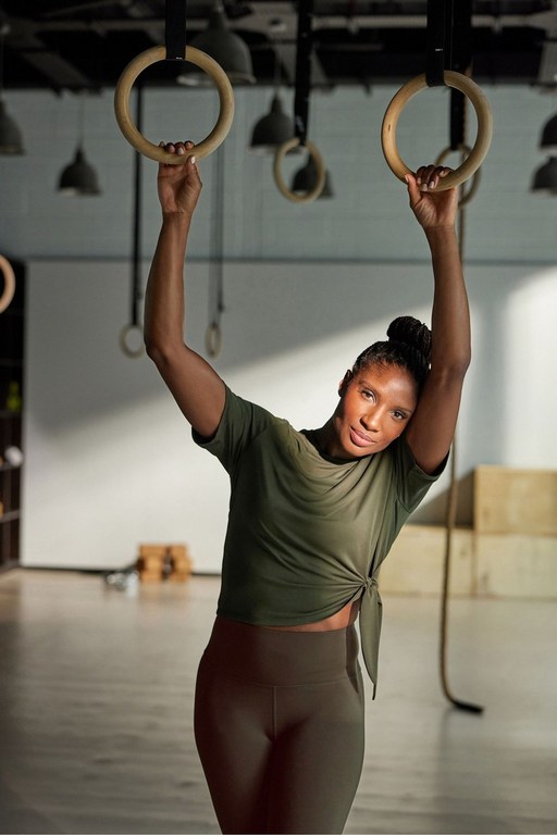 Athletic woman works out on gymnastic rings at an indoor gym. She wears leggings and a tied olive-green t-shirt, looking at the camera.