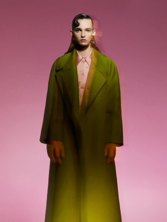 Model wearing an olive green coat and pink shirt against a pink background with a motion blur effect