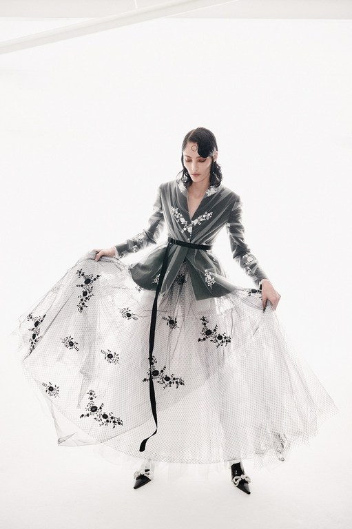 Woman in grey jacket and long white tulle skirt with black floral embellishments poses in a white photo studio with soft lighting.