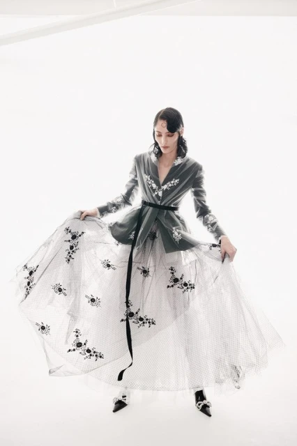 Woman in grey jacket and long white tulle skirt with black floral embellishments poses in a white photo studio with soft lighting.