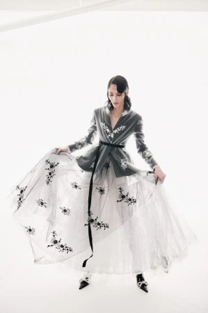 Woman in grey jacket and long white tulle skirt with black floral embellishments poses in a white photo studio with soft lighting.