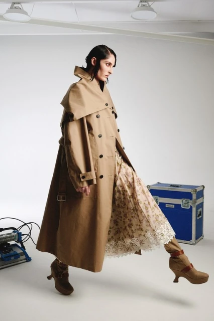 Woman wearing a long tan trench coat over a floral dress and high-heeled boots, striking a pose in a bright photo studio.