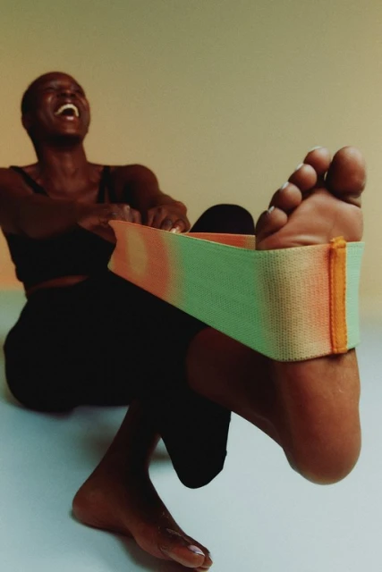 Woman laughs while stretching with a gradient resistance band around her foot, indoors with a light-toned background.