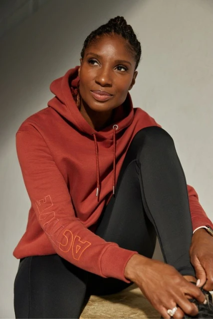 Woman in a rust-colored hooded sweatshirt with black leggings, seated indoors with natural light. She is looking to the side.