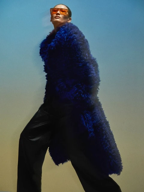 A person strides confidently wearing a blue faux fur coat and glasses, set against a diffused yellow-to-blue gradient background.
