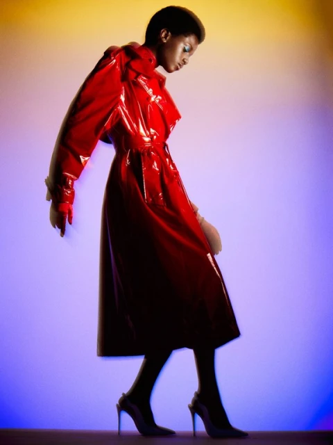 Woman in red patent leather coat walking on illuminated stage, fashion editorial photography with vibrant purple and yellow gradient background.