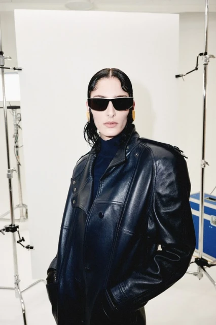 Portrait of a woman with sunglasses and a dark leather coat posing in a studio with white background and light stands.
